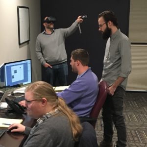One person wearing VR headset in the background, while three men work on computers 