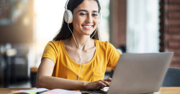 Smiling young woman with headset studying online, using laptop at cafe, taking notes